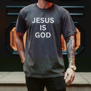 May include: Dark gray t-shirt with the white text "JESUS IS GOD". The shirt has short sleeves and a crew neck. The image shows a person wearing the shirt.
