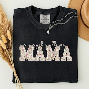 May include: A black short-sleeved t-shirt featuring the white text "an angel calls me" above the word "MAMA" in large block letters. The letters of "MAMA" are filled with a colorful floral pattern. A silver star-shaped necklace rests on the collar.