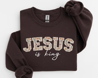 May include: A dark brown sweatshirt with the words "JESUS" and "is king" printed on the front. The word "JESUS" is filled with a floral pattern. The sweatshirt has a crew neck and long sleeves.