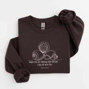 May include: A dark brown sweatshirt with a white seashell design and the text "WHEN YOU GO THROUGH DEEP WATERS I WILL BE WITH YOU ISAIAH 43:2". The sweatshirt has a crew neck and long sleeves.