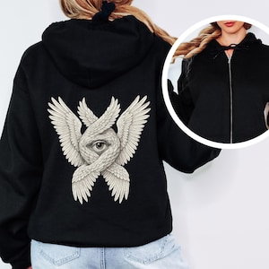 May include: Black zip-up hoodie featuring a detailed cream-colored illustration on the back. The design depicts a central eye surrounded by intertwined wings. The hoodie has a drawstring hood and a full-length zipper.