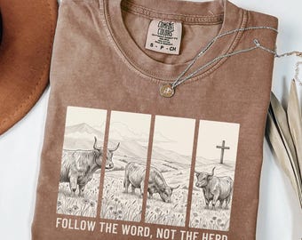 May include: A brown Comfort Colors t-shirt featuring a graphic of three Highland cows in a field with a cross in the background. The text "FOLLOW THE WORD, NOT THE HERD" and "Isaiah 8:11-13" are printed below the graphic. A silver necklace is draped across the shirt.