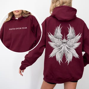 May include: Burgundy hoodie with the text "FAITH OVER FEAR" on the front. The back features a detailed white graphic of multiple wings and eyes. The hoodie has a hood and long sleeves.