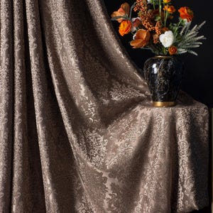 Op de afbeelding: Bruine damast gordijnen met een bloemmotief. Een vaas met oranje, witte en groene bloemen staat op een tafel bedekt met bijpassende stof. De gordijnen en het tafelkleed hebben een glanzende glans.