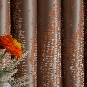 Op de afbeelding: Close-up van bruin en oranje gordijnen met een patroon. De gordijnen hebben een textuur met verticale plooien. Een oranje bloem en een witte bloem zijn zichtbaar in de linker benedenhoek.