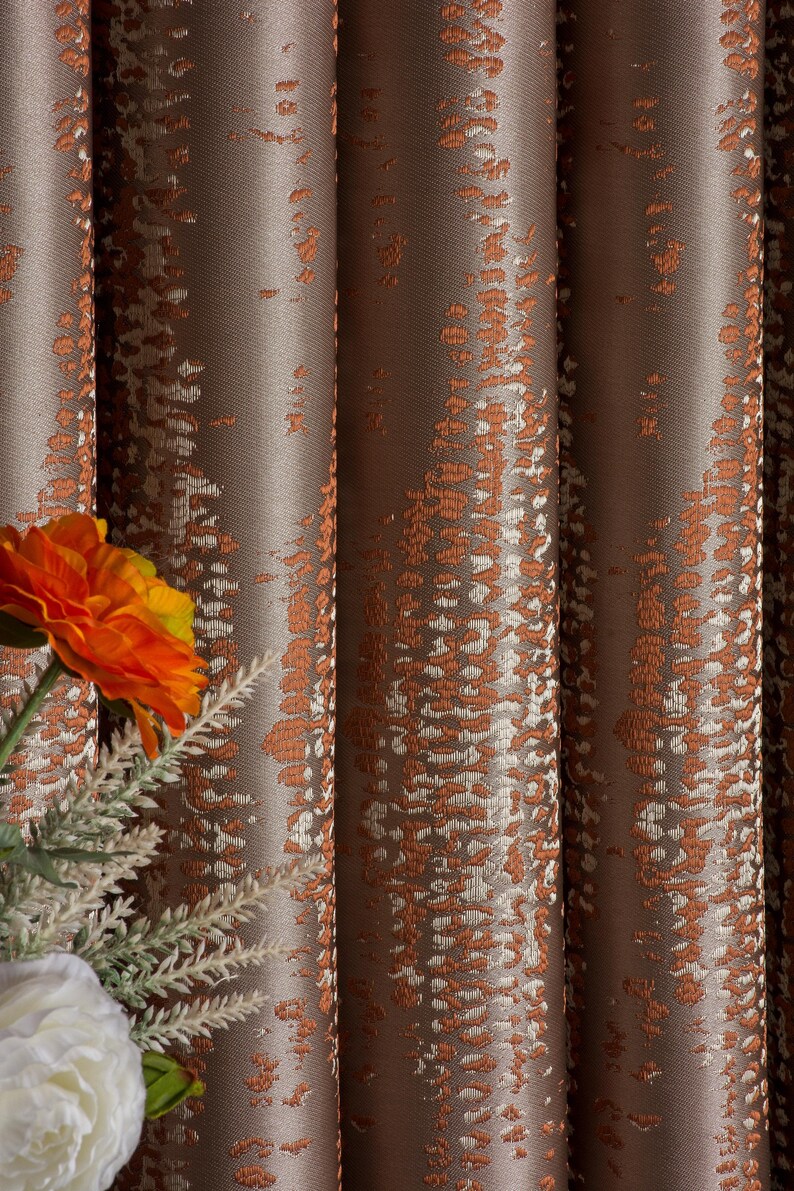 Op de afbeelding: Close-up van bruin en oranje gordijnen met een patroon. De gordijnen hebben een textuur met verticale plooien. Een oranje bloem en een witte bloem zijn zichtbaar in de linker benedenhoek.