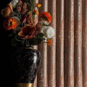 Op de afbeelding: Een zwarte marmeren vaas met gouden accenten bevat een levendig bloemstuk van oranje, witte en bruine bloemen. De vaas staat op een goudkleurige tafel, met getextureerde perzikkleurige gordijnen op de achtergrond.