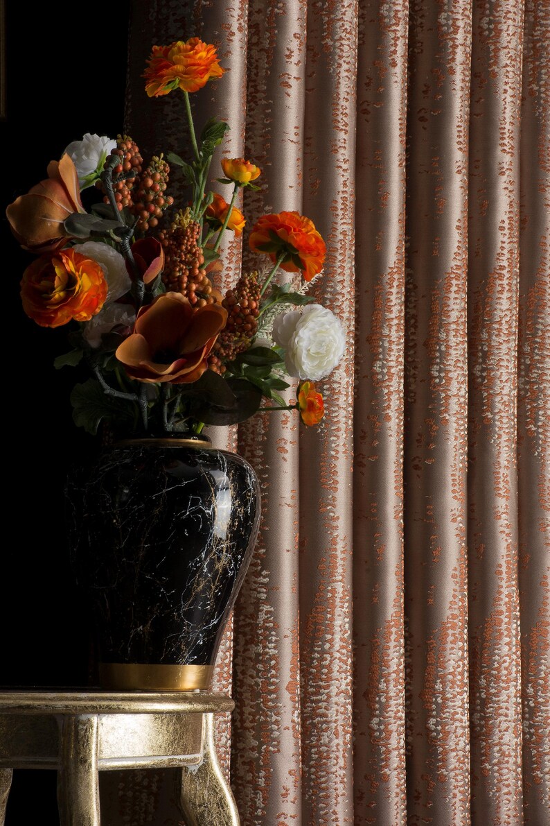 Op de afbeelding: Een zwarte marmeren vaas met gouden accenten bevat een levendig bloemstuk van oranje, witte en bruine bloemen. De vaas staat op een goudkleurige tafel, met getextureerde perzikkleurige gordijnen op de achtergrond.