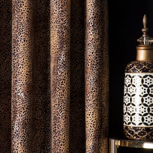 Op de afbeelding: Gordijnen met een luipaardprint in bruin- en beigetinten. Een decoratieve pot met een gouden deksel en geometrisch ontwerp staat op een goudomrandde tafel. De achtergrond is zwart.
