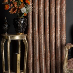 Op de afbeelding: Interieur met een gouden bijzettafel, een zwart-gouden vaas met oranje en witte bloemen en bruine gordijnen met patronen. Op de tafel staat ook een kleine zwart-gouden decoratieve pot.