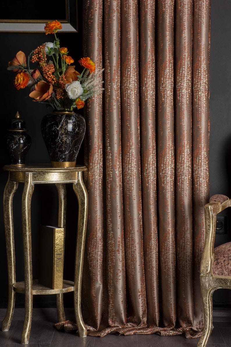 Op de afbeelding: Interieur met een gouden bijzettafel, een zwart-gouden vaas met oranje en witte bloemen en bruine gordijnen met patronen. Op de tafel staat ook een kleine zwart-gouden decoratieve pot.