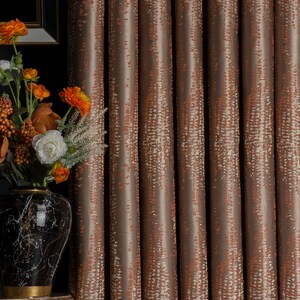 Op de afbeelding: Bruin en oranje gordijnen met een patroon en verticaal ontwerp. De gordijnen hebben een textuur en hangen voor een donkere achtergrond. Links van de gordijnen staat een vaas met bloemen.