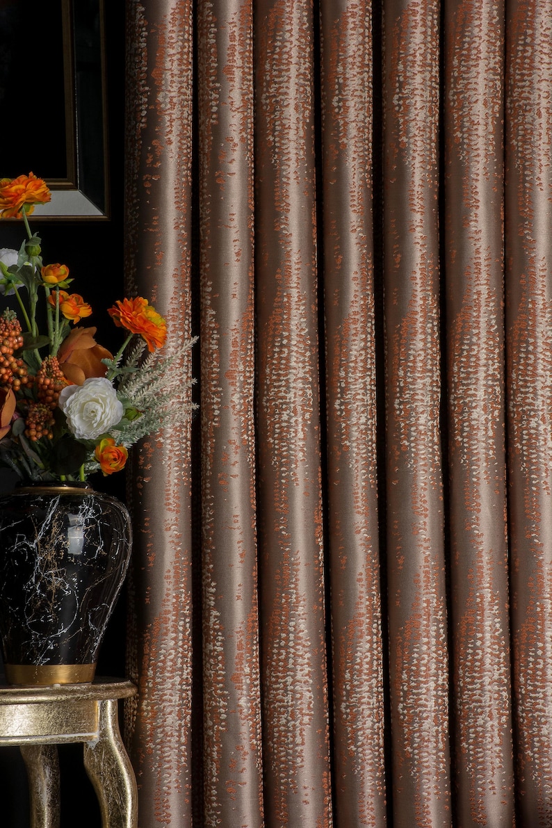 Op de afbeelding: Bruin en oranje gordijnen met een patroon en verticaal ontwerp. De gordijnen hebben een textuur en hangen voor een donkere achtergrond. Links van de gordijnen staat een vaas met bloemen.