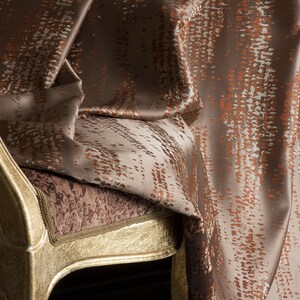 Op de afbeelding: Close-up van een luxe stof gedrapeerd over een goud geverfde stoel. De stof is een mix van bruin, beige en kopertinten, met een textuurpatroon. Het zitkussen van de stoel heeft een patroon en de stoel heeft een gouden rand.