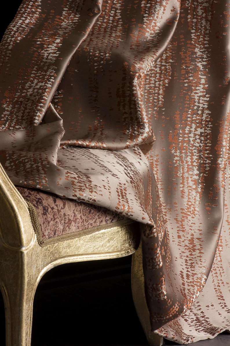 Op de afbeelding: Close-up van een luxe stof gedrapeerd over een goud geverfde stoel. De stof is een mix van bruin, beige en kopertinten, met een textuurpatroon. Het zitkussen van de stoel heeft een patroon en de stoel heeft een gouden rand.