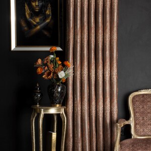 Op de afbeelding: Interieur met een ingelijst portret, een gouden bijzettafel met een vaas bloemen en een gordijn met patroon. Het gordijn heeft een warme bruine kleur met een textuurontwerp. Een decoratieve stoel is ook zichtbaar.