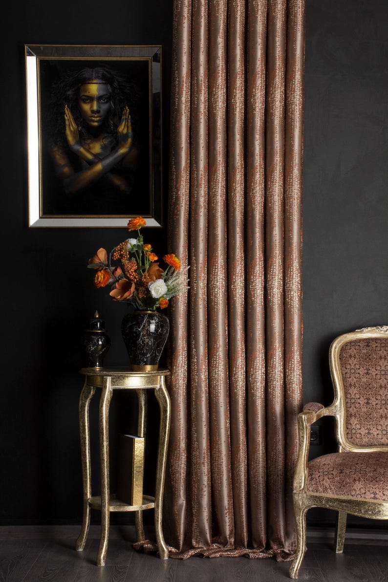 Op de afbeelding: Interieur met een ingelijst portret, een gouden bijzettafel met een vaas bloemen en een gordijn met patroon. Het gordijn heeft een warme bruine kleur met een textuurontwerp. Een decoratieve stoel is ook zichtbaar.