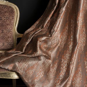 Op de afbeelding: Een luxe bruin gordijn met een textuurpatroon van oranje en witte verticale lijnen. De stof drapeert elegant, met een glimp van een goudomrandde stoel met een patroon op de rug op de achtergrond. Het gordijn voegt elegantie toe.