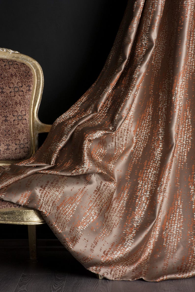 Op de afbeelding: Een luxe bruin gordijn met een textuurpatroon van oranje en witte verticale lijnen. De stof drapeert elegant, met een glimp van een goudomrandde stoel met een patroon op de rug op de achtergrond. Het gordijn voegt elegantie toe.