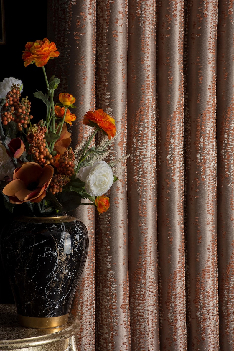 Op de afbeelding: Een decoratief arrangement met een zwarte marmeren vaas met gouden accenten, gevuld met oranje en witte bloemen. De achtergrond toont getextureerde, bruine gordijnen met een subtiel patroon.