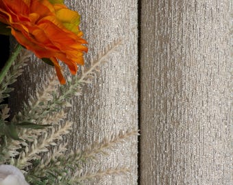 May include: Close-up of two rolls of textured, light beige fabric with subtle silver flecks. An orange and yellow flower, white flower, and sprigs of greenery are in the foreground, adding a touch of color and contrast to the neutral background.