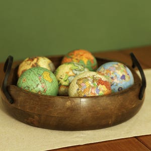 May include: A wooden bowl containing several miniature globes. The globes display detailed world maps in various colours, including green, orange, and blue. The bowl is dark brown with black handles, resting on a beige surface.