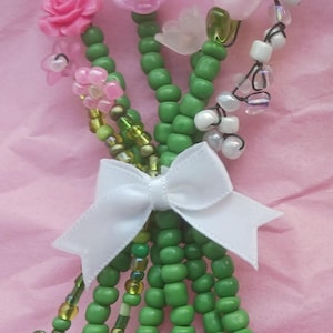 Op de afbeelding: Een kralenboeket met groene stelen en diverse bloemmotieven. De bloemen omvatten roze rozen, doorschijnende bloemblaadjes en witte kralenclusters. Een witte strik zet de stelen vast. De achtergrond is roze.