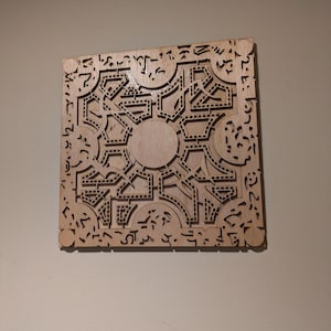 May include: A square, wooden puzzle box with intricate geometric patterns and symbols carved into its surface. The box features a central circular design and is made of light-colored wood. The overall design is symmetrical and detailed.