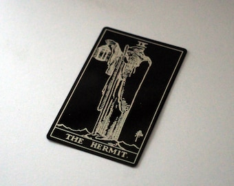 Puede incluir: Carta de tarot negra con una ilustración blanca del Ermitaño. El Ermitaño es una figura alta con una linterna y un bastón, de pie en la cima de una montaña. La carta dice "THE HERMIT".