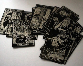 May include: A collection of black tarot cards with white illustrations and text. The cards are fanned out, displaying various images and titles such as "The Fool," "The Moon," and "The Sun." The cards are rectangular and have a vintage aesthetic.