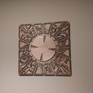 May include: A wooden puzzle box with intricate laser-cut designs. The box is square with a circular center and detailed patterns around the edges. The wood has a natural, light brown color.