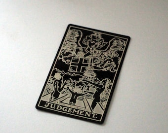 Puede incluir: Carta de tarot en blanco y negro con la palabra "JUICIO" en la parte inferior. La carta representa un ángel con las alas extendidas, tocando una trompeta, y una cruz arriba. Cuatro figuras se levantan del suelo.