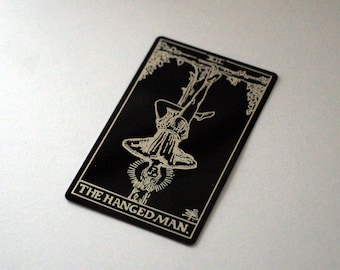 Puede incluir: Una carta de tarot negra con una ilustración blanca de una persona colgada boca abajo de una rama de árbol. La carta dice "THE HANGED MAN."