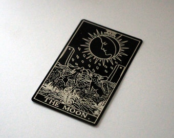Puede incluir: Una carta de tarot negra con una ilustración detallada de la luna, dos perros y agua. La carta tiene un borde blanco y las palabras "THE MOON" en la parte inferior.