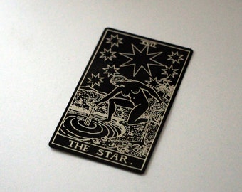 Puede incluir: Carta de tarot en blanco y negro con la imagen de una mujer vertiendo agua de una jarra en una piscina. La carta se titula "La Estrella" y tiene el número romano "XVII" en la esquina superior derecha.