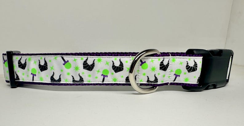 Op de afbeelding: Een witte hondenriem met een paarse rand en een herhalend patroon van groene appels en zwarte silhouetten van een kroon. De riem heeft een zilveren D-ring en een zwarte plastic gesp.