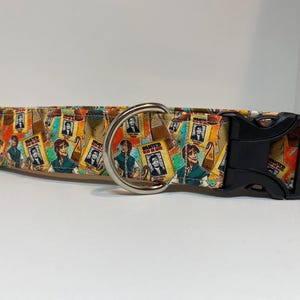 May include: A colorful dog collar with a black buckle and silver D-ring. The collar features a repeating pattern of wanted posters with cartoon images of a man with brown hair and a blue shirt.