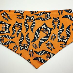 May include: Orange dog bandana with a repeating pattern of a character's face in white, navy, and black. The bandana is a triangle shape, suitable for pets. The design is a cartoon-style illustration.