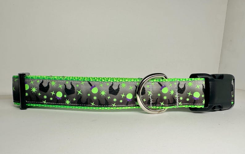 Op de afbeelding: Een groene en grijze hondenriem met een zwarte gesp en een zilveren ring. De riem heeft een herhalend patroon van een heksensilhouet, een ketel en sterren.