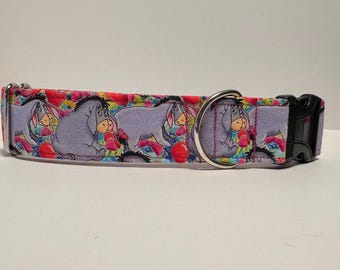 May include: A colorful dog collar with a repeating pattern of Eeyore from Winnie the Pooh. The collar is made of fabric and has a silver metal buckle and a black plastic clasp.