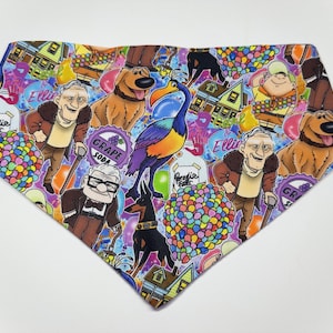 May include: A colorful dog bandana featuring characters from the animated movie Up. The bandana has a blue background with images of Carl Fredricksen, Russell, Dug, Kevin the bird, and balloons. The bandana also has the text "Ellie" and "Grape Soda".