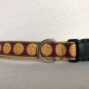 May include: A purple pet collar featuring a repeating pattern of gold circular icons with a lightning bolt design. The collar includes a black plastic side-release buckle and a silver-toned metal D-ring for attaching a leash or identification tags.
