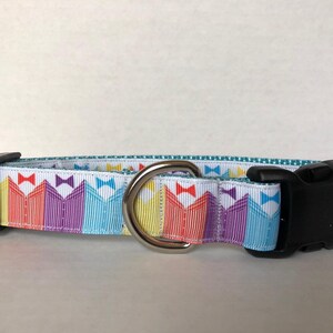 May include: A white dog collar with a colorful pattern of stripes and bowties. The collar has a silver D-ring and a black plastic buckle.