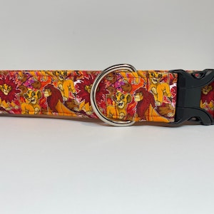 May include: A colorful dog collar with a repeating pattern of lions from the movie The Lion King. The collar is orange, red, and yellow with a black buckle and silver D-ring.