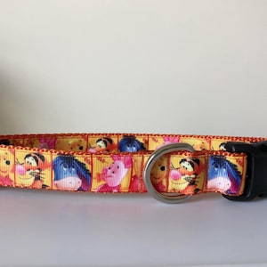 May include: A yellow, orange, and pink dog collar with a black buckle and a silver ring. The collar features a repeating pattern of Winnie the Pooh and his friends.