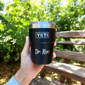 May include: A black Yeti tumbler with a silver rim and lid, held against a blurred green background. The Yeti logo is displayed on the front, with the name "Dr. Ron" in a cursive font below it. The tumbler is designed to keep beverages cold or hot.