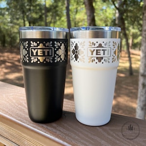 May include: Two stainless steel tumblers, one matte black and one matte white, each featuring a metallic silver band near the top rim with a repeating floral pattern and the YETI brand name centered in a rectangular frame on the front.