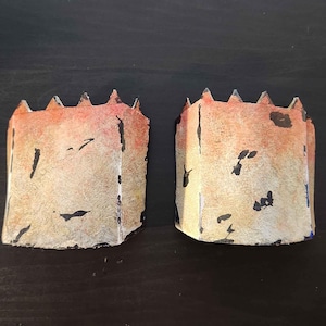 Könnte beinhalten: Zwei kleine, strukturierte, beigefarbene Papiermaché-Türme mit schwarzen und roten Akzenten. Die Türme haben ein gezacktes, schlossartiges Oberteil.