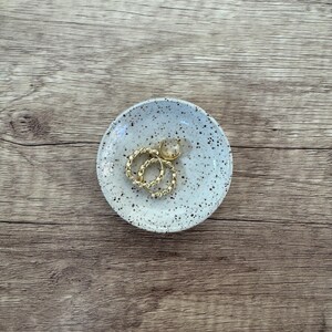 May include: A white ceramic trinket dish with brown speckles. The dish has two gold rings and a gold bead inside.