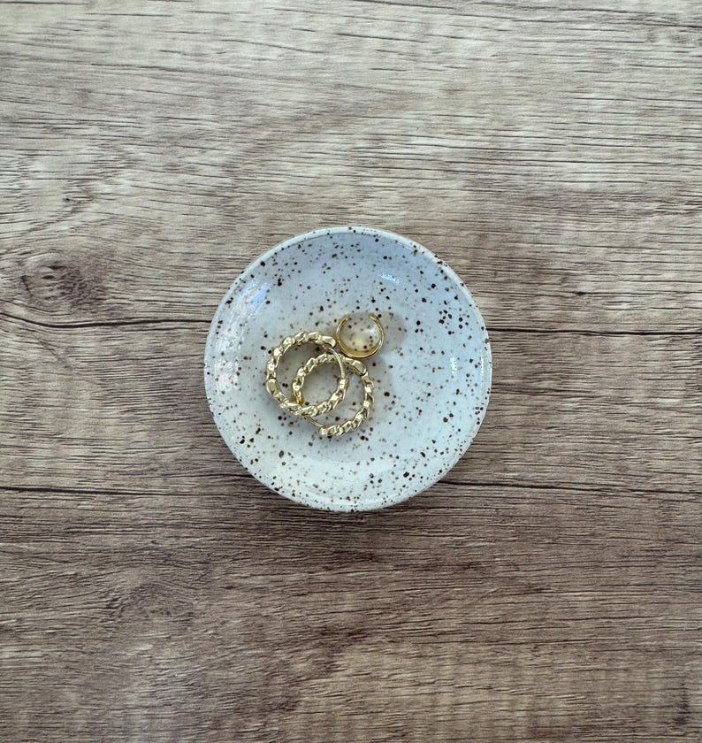 May include: A white ceramic trinket dish with brown speckles. The dish has two gold rings and a gold bead inside.